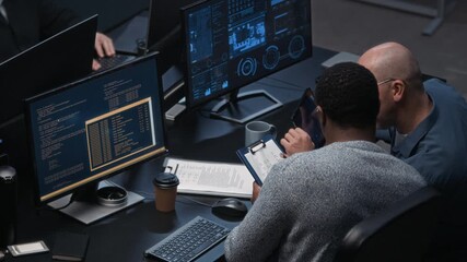 Two diverse male cybersecurity workers analyzing documents and monitoring digital data on computer screens in dark office, collaborating on network security tasks - Powered by Adobe