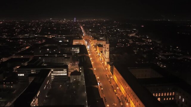 Luftaufnahme von Muenchen, Deutschland bei Nacht mit Ludwigstrasse, St Ludwig Kirche, Siegestor, beleuchteten Strassen, Nachtverkehr, Fussgaengern und Panorama der Innenstadt.