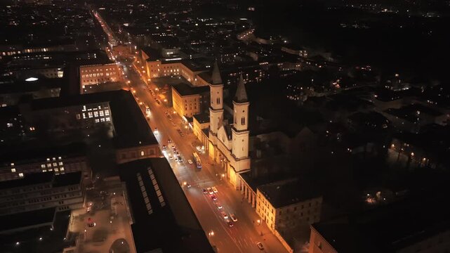 Night drone footage over Munich, Germany Innenstadt featuring Ludwigstrasse boulevard, St Ludwig Church, Siegestor triumphal arch, street lighting, moving cars, and pedestrian activity.