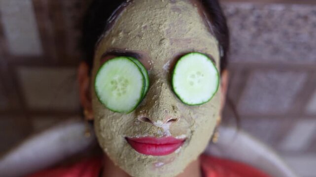 A beautiful woman with facial clay mask on face and fresh cucumber on eyes relaxing at Home. Portrait of beautiful spa girl. Beauty, healthcare. Multani Mitti and green Face Pack. Asian Indian lady