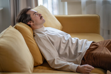 Thoughtful tired man lying on sofa at home, looking upward in self reflection. Pensive fatigued male rest on couch in living room, lost in thoughts after long day. Decision making, loneliness, burnout