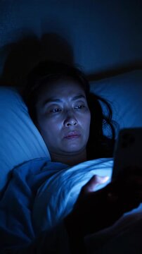 A person lying in bed, illuminated by the blue light of a phone screen, reflects a moment of nighttime solitude and digital engagement in a dark room.
