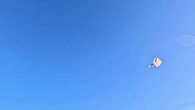 Small paper airplane glides through clear blue sky, drifting farther from camera as it tilts and catches wind; minimal scene suggests freedom, travel and imagination with copy space.