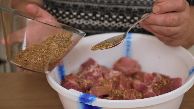 Sprinkling spice mix over raw pork pieces for marinade. Adding seasoning to fresh meat for barbecue preparation. Mixing herbs and ground spices with pork chunks for grilling process