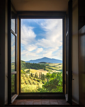 View of golden fields meet rolling hills under a bright sky, framed by an open window revealing the idyllic landscape, Montepulciano, Tuscany, Italy.