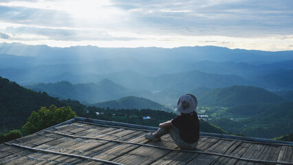 Obraz premium Rear view of a woman sitting on wooden terrace and looking at a mountain view before sunset