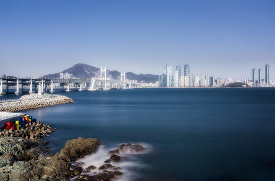 View of the vast, still ocean reflects the towering skyline under a bright sky, with a distant bridge connecting to the shore, Busan, Busan, South Korea.