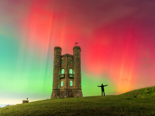 View of a silhouette of a person with arms outstretched against a vibrant aurora-filled sky, standing near a stone tower on a grassy hill, Broadway Tower, The Cotswolds, United Kingdom.
