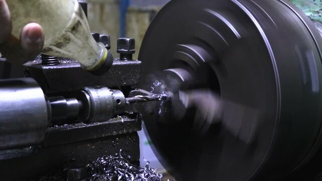 Close-up of a drilling process on a lathe machine. The turner's hands are visible applying lubricant as the drill bit enters the rotating metal part, creating shavings in an industrial workshop.