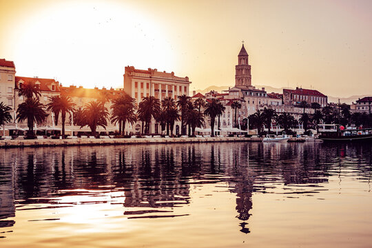 View of the warm glow of sunset casting long reflections on the water in front of the historic buildings and palm trees, Split, Split-Dalmatia County, Croatia.