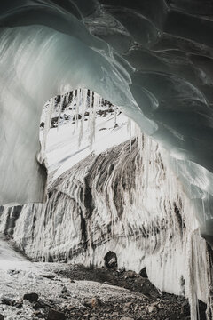 View of a glacial cavern with dripping icicles framing a snow-covered mountain, icy textures, and rugged terrain, creating a cold, awe-inspiring scene, Pontresina, Grisons Canton, Switzerland.