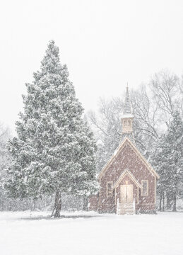 View of a quaint red chapel stands nestled amidst snow-laden trees, its warm light a beacon in the serene, wintry landscape,Yosemite Valley, California, United States.