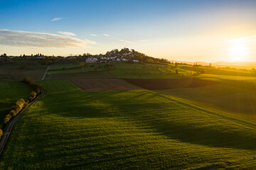 Aerial view of golden sunlight bathes the lush green fields and small village atop a hill, casting long shadows across the landscape, Otzberg, Hessen, Germany.