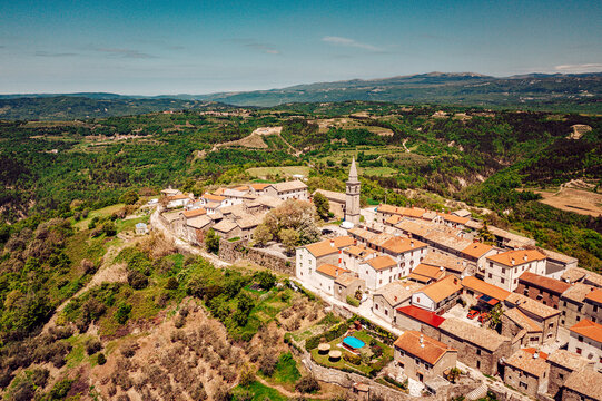Aerial view of terracotta rooftops cascading down the hillside, a church steeple piercing the skyline amidst the verdant landscape, Primosten, Sibenik-Knin County, Croatia.