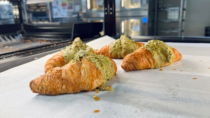 Golden croissants with pistachio filling cooling on parchment paper in a bakery workspace. Delicious homemade pastry, fresh dessert preparation and food photography background.
