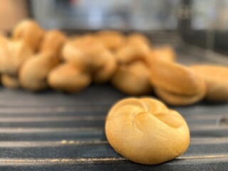 Close-up of a small pretzel-shaped bread roll in focus, with a pile of fresh bakery buns blurred in the background, ideal for food photography.