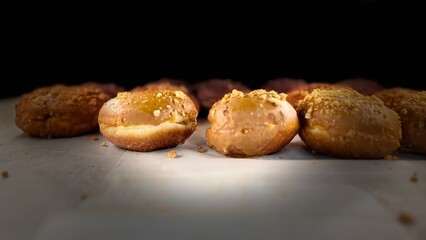 Glazed donuts with nut topping arranged in a row on baking paper. Freshly made filled doughnuts with shiny icing, soft texture, and dark moody background. Sweet bakery dessert, close-up food photograp
