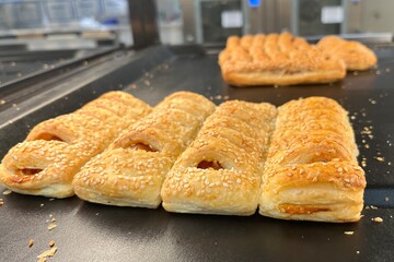 Fresh baked puff pastry rolls with sesame seeds on a bakery tray. Golden flaky pastries displayed in a shop, close-up food background with shallow depth of field.