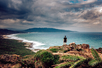 View of a lone figure standing atop rocky cliffs overlooking the wavy, turquoise sea under a moody sky, Taipei City, Taipei City, Taiwan.