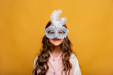 Overjoyed little girl wearing festive masquerade mask and pink sweatshirt on yellow background. Child having fun at carnival party in Brazil or Venice. Carnival promotion and holidays concept