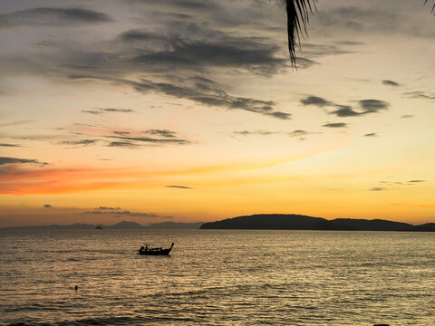 View of a silhouetted longtail boat glides across the tranquil sea under a vibrant sunset sky painted with orange and grey hues, Ao Nang, Krabi, Thailand.