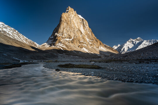 View of a majestic, sun-kissed mountain peak rising sharply above a glacial river valley, casting long shadows across the rocky terrain, Zanskar, Ladakh, India.