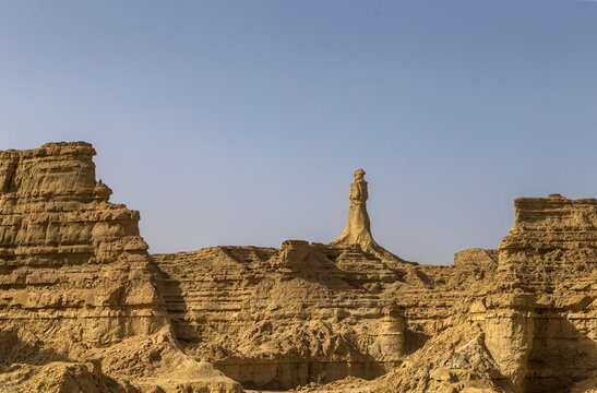 View of rugged, sun-baked cliffs and a solitary, spire-like rock formation pierce the serene sky, creating a striking contrast, Makran Coastal Highway, Balochistan, Pakistan.
