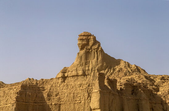 View of a weathered, golden-brown rock formation standing tall against the clear blue sky, its sculpted facade reminiscent of an ancient sentinel, Makran Coastal Highway, Balochistan, Pakistan.