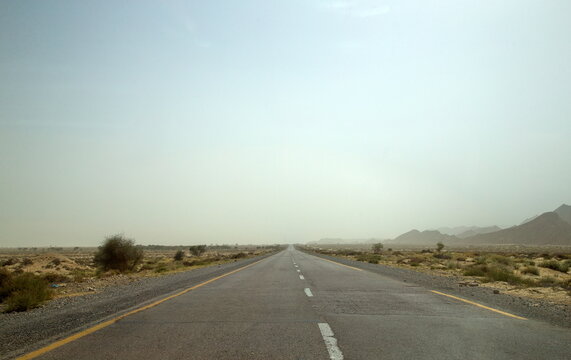 View of a long, straight highway cutting through the arid landscape under a hazy sky, with sparse vegetation and distant mountains, Makran Coastal Highway, Balochistan, Pakistan.