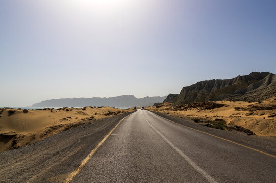 View of asphalt ribbon winding through sun-baked desert, meeting distant mountains under a vast sky, a stark contrast of terrain, Makran Coastal Highway, Balochistan, Pakistan.