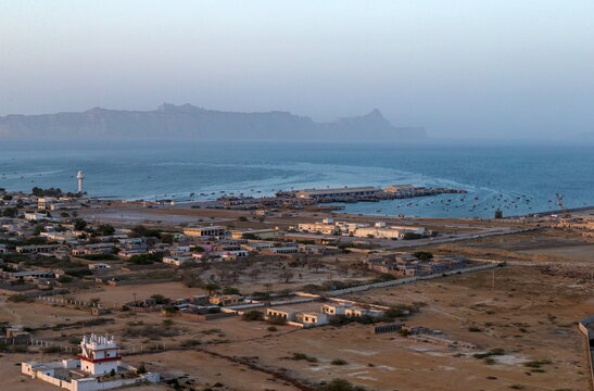 View of the arid landscape meeting the tranquil sea, where modest buildings nestle under a vast sky, as mountains fade into the horizon, Gwadar, Balochistan, Pakistan.
