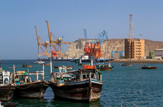 View of boats bobbing in the harbor, with industrial cranes casting long shadows against the backdrop of rocky cliffs, Gwadar, Balochistan, Pakistan.