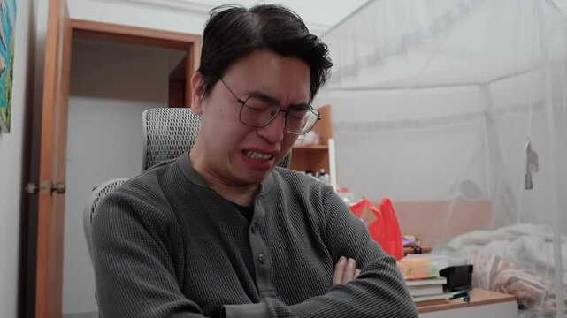 A young Asian Chinese man is crying uncontrollably in his bedroom at his computer desk because of heartbreak