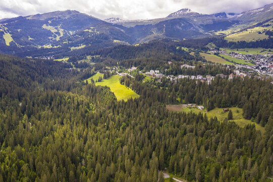 Aerial view of dense evergreen forests meeting the village nestled in the valley, with snow-capped mountains in the distance, Flims, Grisons, Switzerland.
