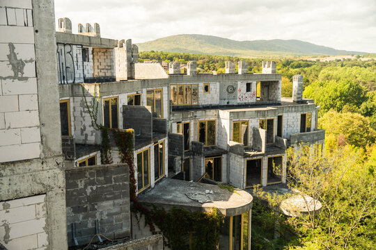 View of an unfinished, crumbling castle-like structure, where concrete contrasts with the encroaching green foliage, under a cloudy sky, Costa del croco, Tsarevo, Bulgaria.