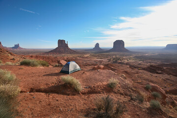 Tent set on red desert ground overlooking iconic sandstone buttes of Monument Valley Navajo Tribal Park, USA. Vast open landscape of the American Southwest under a clear blue sky.