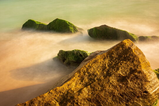 View of moss-covered rocks kissed by the soft, blurred caress of the ocean's edge under a serene sky, Gawadar, Balochistan, Pakistan.