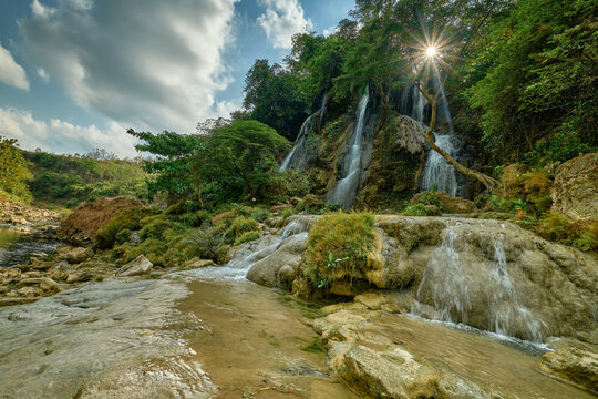 View of cascading waterfalls plunge down moss-covered rocks amidst lush greenery, sunbeams piercing the canopy above, Sri Gethuk Waterfall, Yogyakarta, Special Region of Yogyakarta, Indonesia.