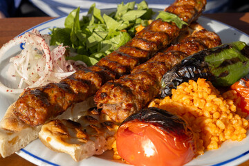 Adana kebab plate with two juicy grilled meat skewers, bulgur pilaf, charred pepper and tomato, fresh salad and onion garnish for an authentic turkish meal