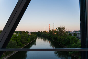 View of calm waters reflecting the soft sky, framed by green trees and distant industrial chimneys, a blend of nature and industry, Vienna, Vienna, Austria.