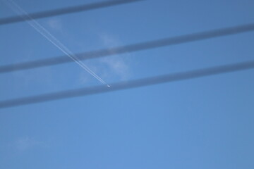 Jet plane flying high leaving white contrail in blue sky behind blurred power lines