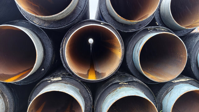 Close‑up of stacked pre‑insulated steel pipes for a district heating network, with large round openings forming an abstract industrial pattern and visible rust layers inside the tubes.