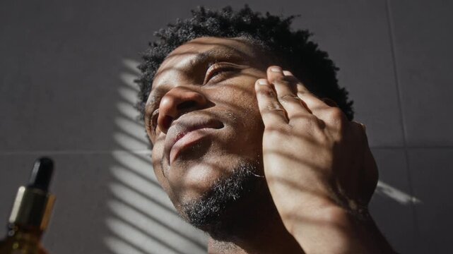 Close-up low angle view of Black young man with beard applying facial serum directly with dropper and rubbing it into cheek with light movements