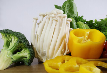 A large cluster of white mushrooms is placed between sliced ​​yellow bell peppers and green broccoli on a table with a white background.

