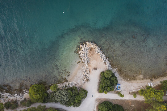 Aerial view of the tranquil turquoise waters meeting the rocky coastline, fringed by verdant trees and pathways, Rovinj, Istria County, Croatia.