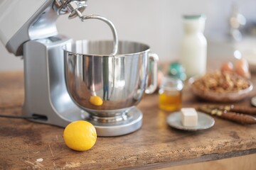 A silver stand mixer sits beside a bright lemon. Butter, honey, and milk rest on a wooden table,...