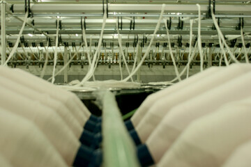 Close-up perspective of white yarn strands being fed through horizontal metal bars and rollers on industrial spinning machinery in a textile factory floor.