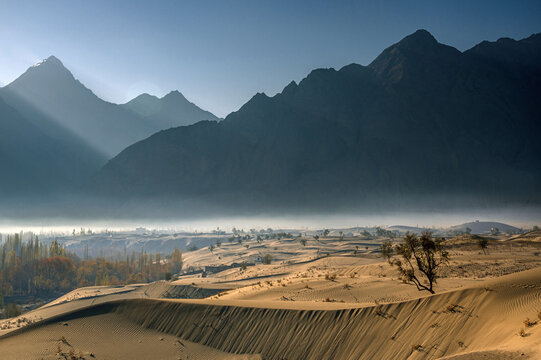 View of sunlight filtering through the misty air over the soft, undulating dunes, contrasting with the stark, dark silhouettes of rugged mountains, Skardu, Gilgit Baltistan, Pakistan.
