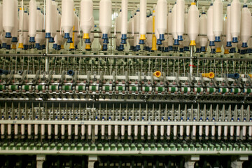 Wide view of multiple rows of white yarn spools hanging vertically above complex spinning machinery in a bright textile mill, highlighting mass industrial production.