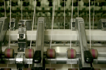 Close-up of industrial textile machinery processing vertical strands of white yarn through a series of metal guides and rollers on a factory floor.